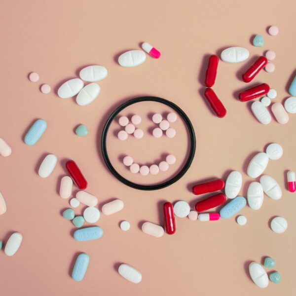 Assorted pills and capsules arranged around and inside a circular dish, forming a smiling face on a pastel background.
