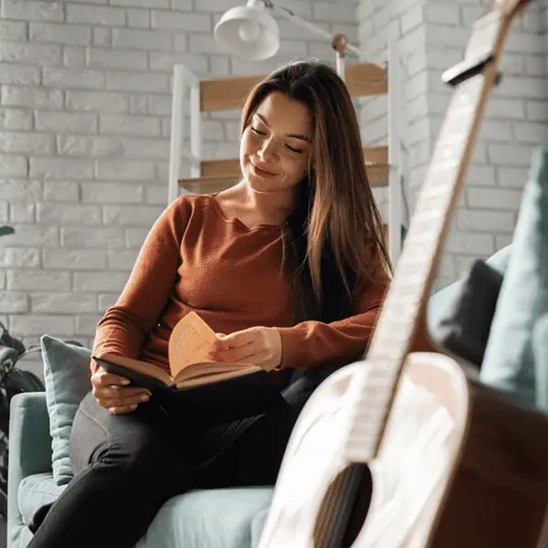 A person sitting on a couch in a bright room, reading a book with a guitar resting nearby.