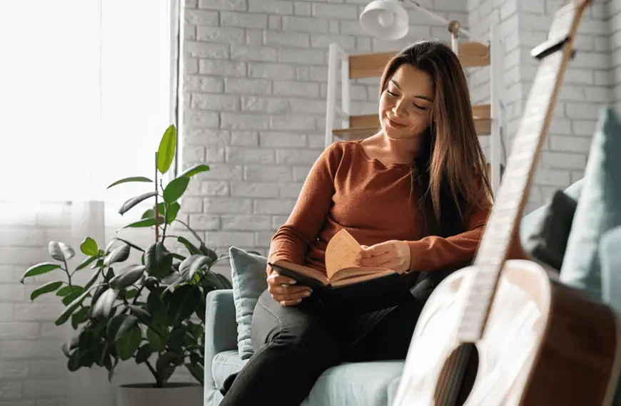 A person sitting on a couch in a bright room, reading a book with a guitar resting nearby.